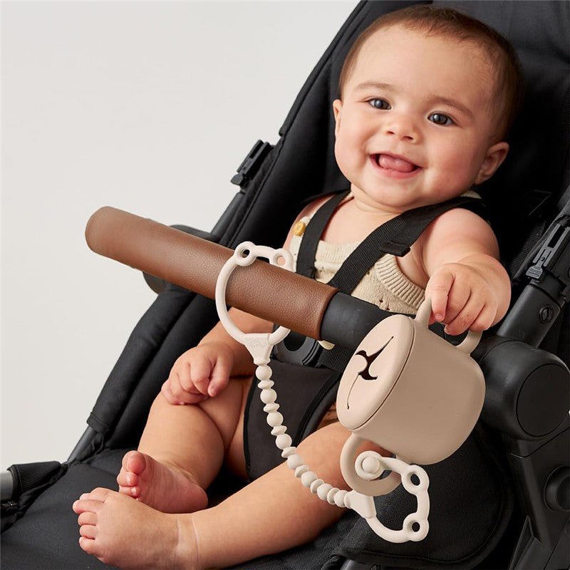 A smiling baby in a stroller holds a silicone snack cup strap with a cup attached, featuring a light tan color and curved design.