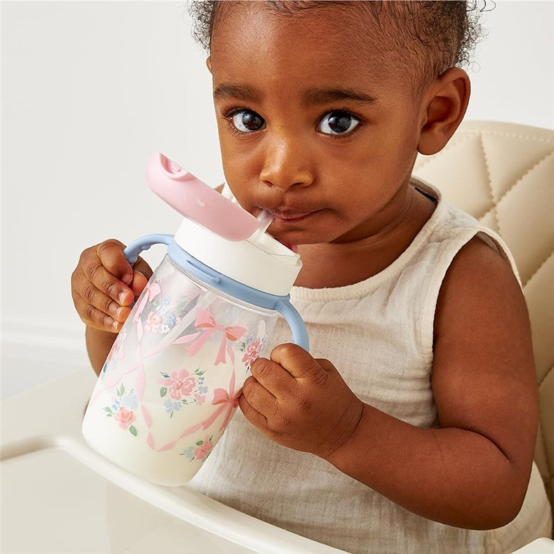 A toddler holds an Itzy Ritzy Straw Sippy Cup featuring a floral design with ribbons, drinking from it while seated.