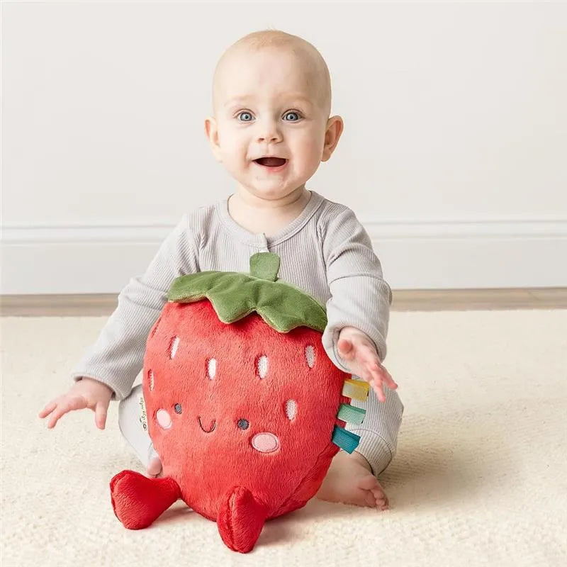 Infant sitting on a light rug, smiling, while holding a plush strawberry toy with colorful tags and a happy face.