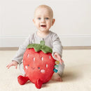 Infant sitting on a light rug, smiling, while holding a plush strawberry toy with colorful tags and a happy face.