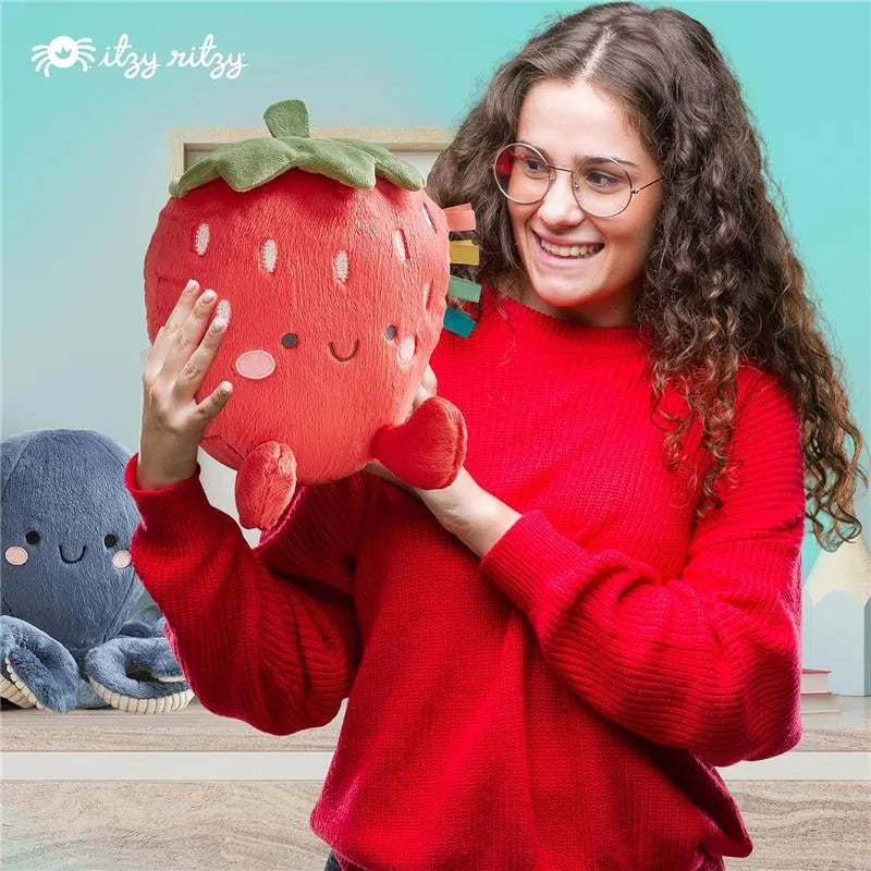 A woman in a red sweater smiles while holding a plush strawberry toy from Itzy Ritzy, with a blue plush octopus in the background.