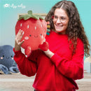 A woman in a red sweater smiles while holding a plush strawberry toy from Itzy Ritzy, with a blue plush octopus in the background.
