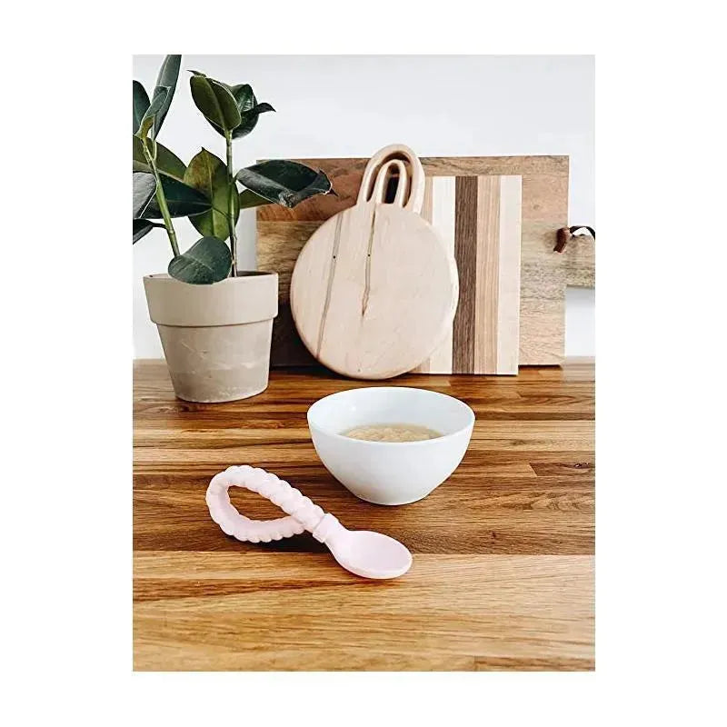Pink Itzy Ritzy Sweetir Spoons silicone baby utensil set displayed next to a bowl of food, a decorative plant, and wooden cutting boards.