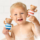 A happy child holds two wind-up bath toys shaped like bears, one in blue stripes and the other in red stripes, in a bubbly bath.