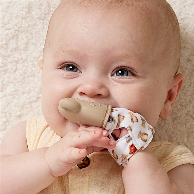 Baby smiling while holding an Itzy Ritzy Teething Mitt with a beige silicone tip and classic bear-patterned fabric.