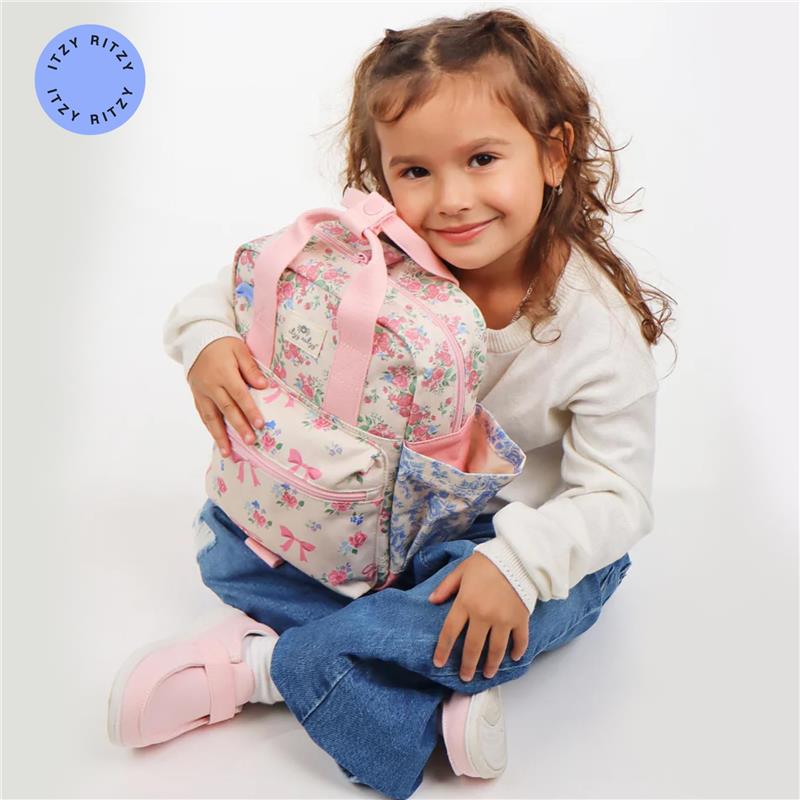 A smiling girl in a white sweater and blue jeans holds an Itzy Ritzy Toddler Backpack featuring floral patterns and pink accents.