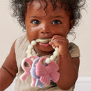 Baby smiling while holding a multi-textured silicone teether featuring colorful bows, safe for chewing and play.