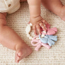 Multi-textured silicone teether bows in pink, blue, and white held by a baby on a textured blanket.