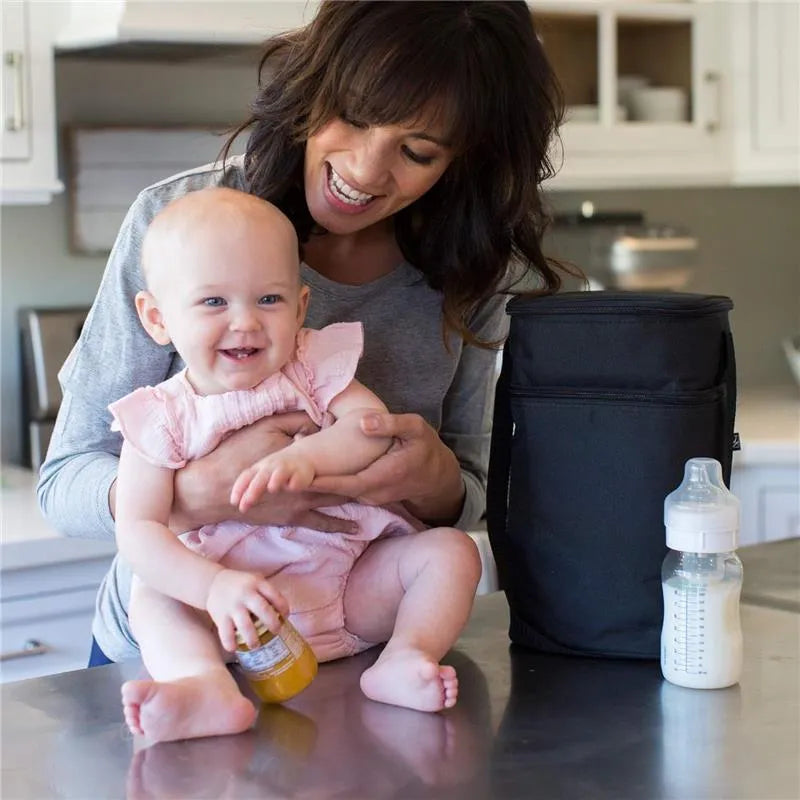 A smiling mother holds her baby in a kitchen, next to a black insulated cooler and a bottle of milk.
