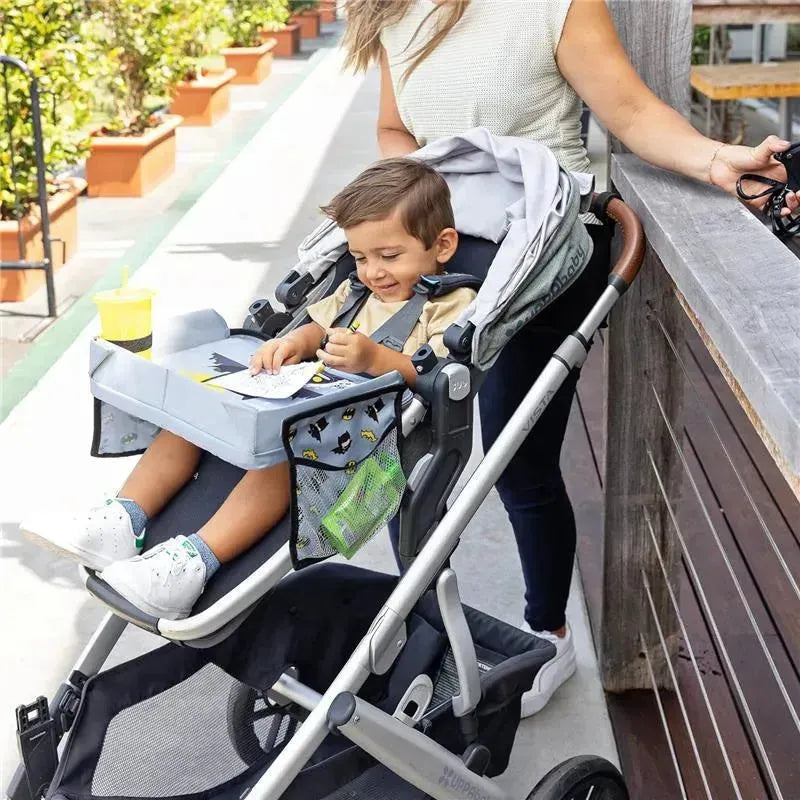 A young boy sits in a stroller, drawing on a Batman-themed travel tray, while a woman holds the stroller.