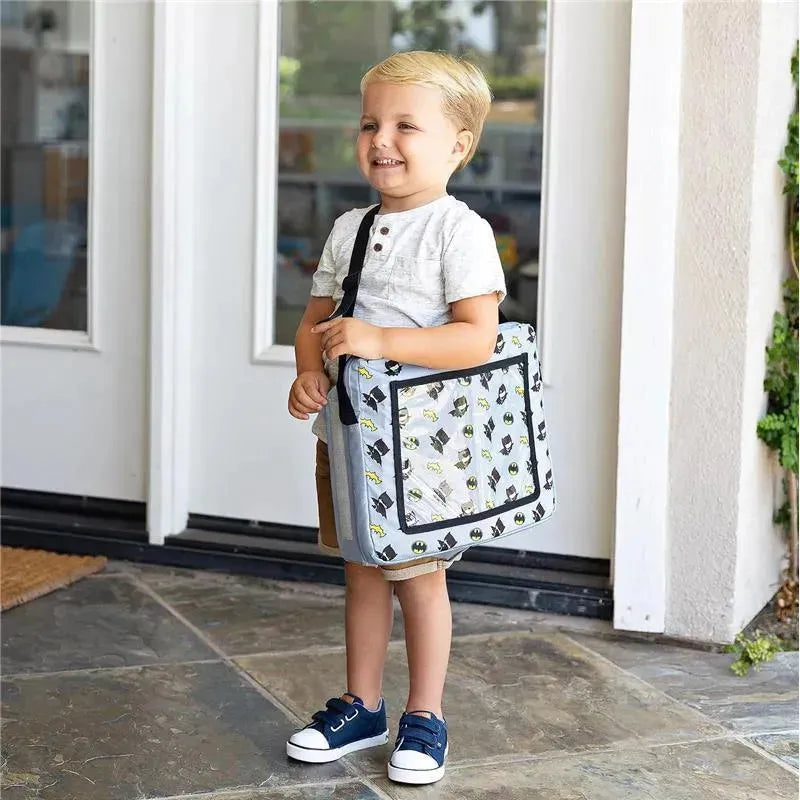 A smiling child holds a DC Comics Batman travel bag, featuring an all-over print of Batman icons, while standing at a doorway.