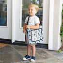 A smiling child holds a DC Comics Batman travel bag, featuring an all-over print of Batman icons, while standing at a doorway.