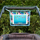 Toddler travel tray with colorful stitching holds a tablet above a stroller, surrounded by greenery.