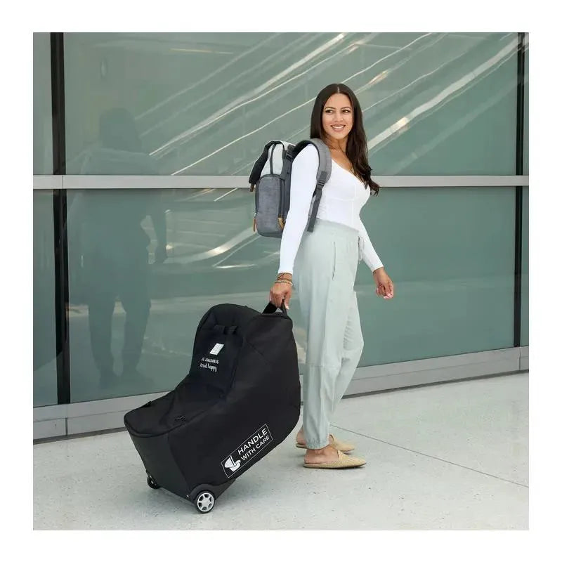 Black J.L. Childress car seat travel bag with wheels, carried by a woman in an airport terminal.
