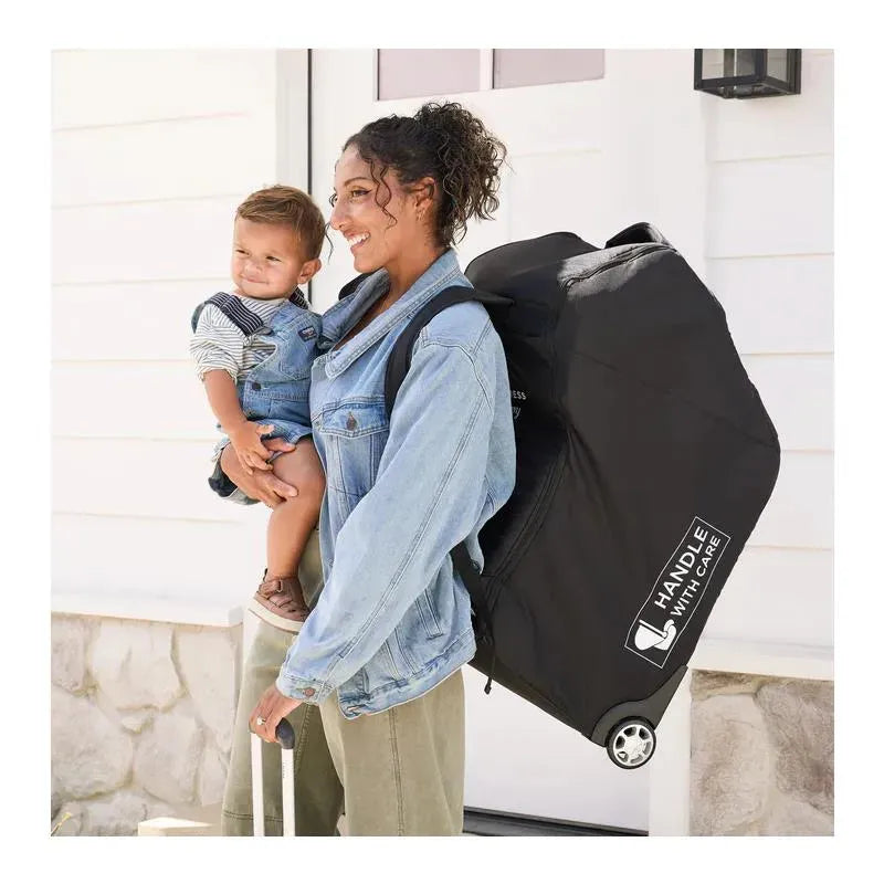 Woman carrying a child on her hip while wheeling a black J.L. Childress infant car seat travel bag with wheels.