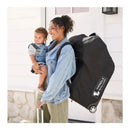 Woman carrying a child on her hip while wheeling a black J.L. Childress infant car seat travel bag with wheels.
