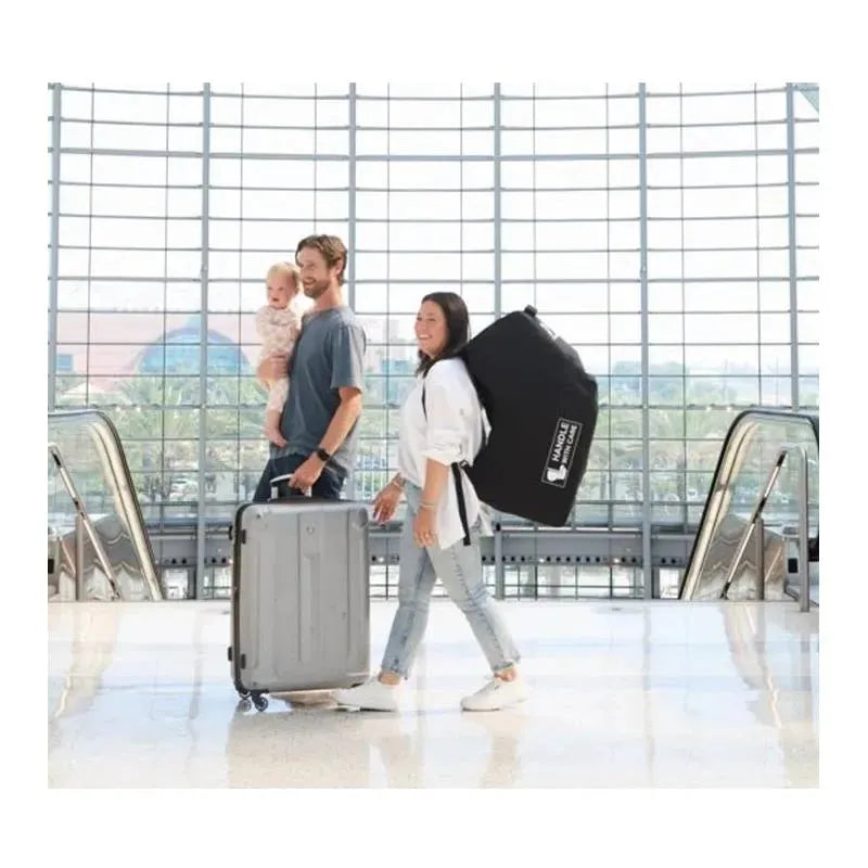 Family at an airport: a man holds a baby while pushing a suitcase, and a woman carries a padded black car seat travel bag.