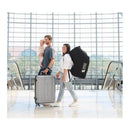 Family at an airport: a man holds a baby while pushing a suitcase, and a woman carries a padded black car seat travel bag.