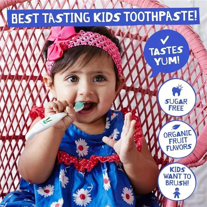 A toddler with a pink headband smiles while holding a toothbrush and showing a tube of raspberry-flavored toothpaste.
