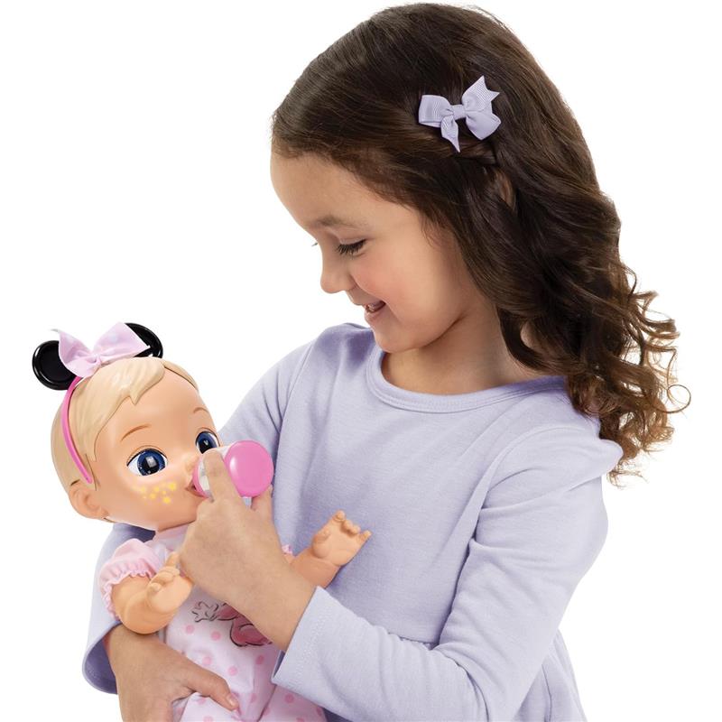 A girl in a lavender top smiles as she feeds a 13-inch interactive baby doll with blonde hair and a pink bottle.