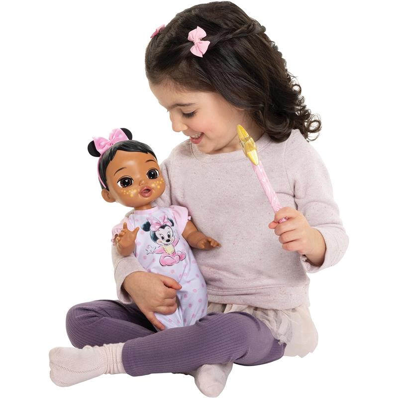 A young girl holds a 13-inch interactive baby doll with brown hair, wearing a pink Minnie Mouse outfit, while smiling and playing.