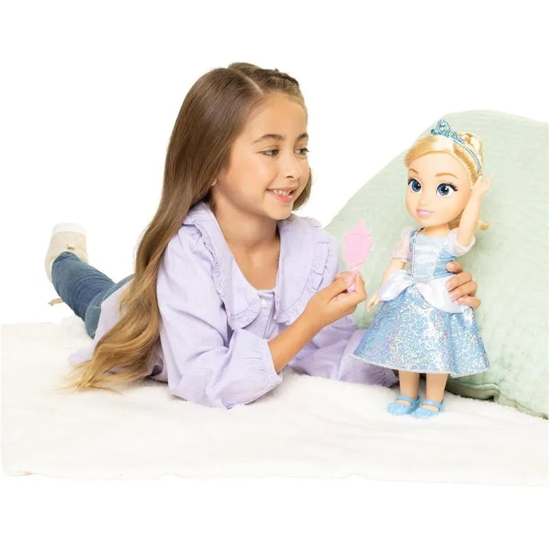 Girl playing with a Cinderella doll, showing a pink accessory, on a soft surface with a green pillow in the background.
