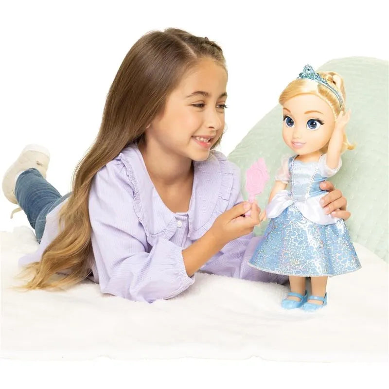 A child smiles while holding a pink wand, playing with a Cinderella large doll wearing a sparkly blue dress.