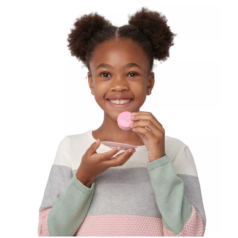 A girl with puffy hair smiles while holding a pink macaron and a teacup. She wears a colorful striped sweater.