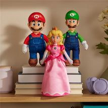 Plush toys of Princess Peach, Mario, and Luigi stand on stacked books, featuring colorful outfits and friendly expressions.