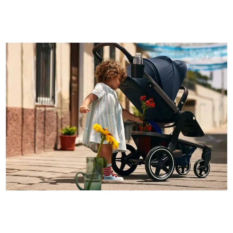 A young child in a striped dress interacts with flowers while standing next to a stroller equipped with a cup holder.