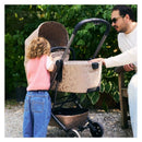 Child in a pink sweater interacts with a Joolz Aer2 Fold Cot in Chic Renaissance, Taupe, while an adult observes nearby.