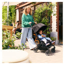 A woman in a green sweater pushes a Joolz Aer2 stroller in Space Black with a child inside, near a cafe.