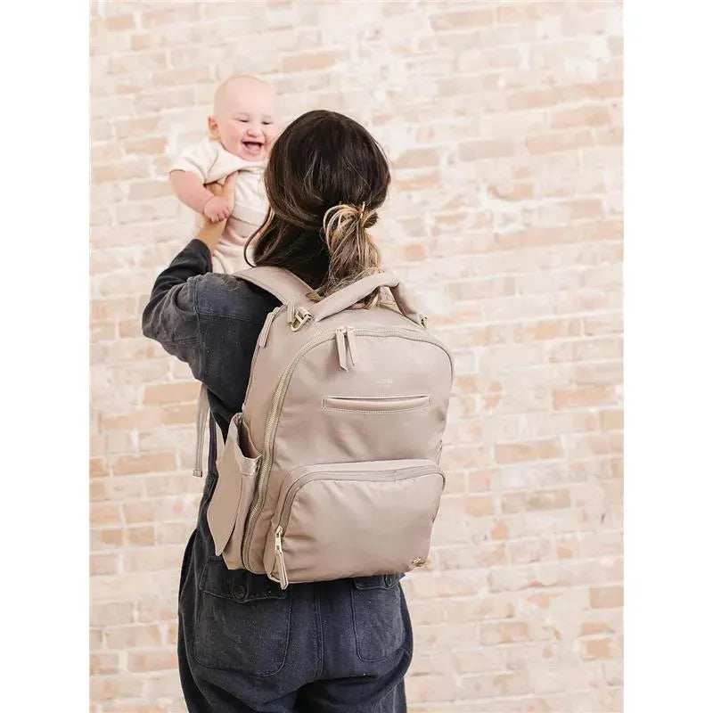 Taupe Jujube Classic Backpack worn by a person holding a smiling baby, set against a light brick wall.