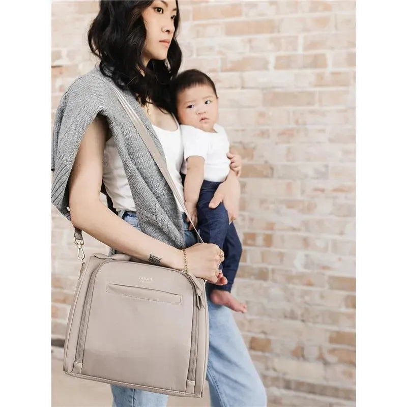 A woman holding a baby while carrying a taupe Jujube insulated bottle bag on her shoulder, against a brick wall backdrop.