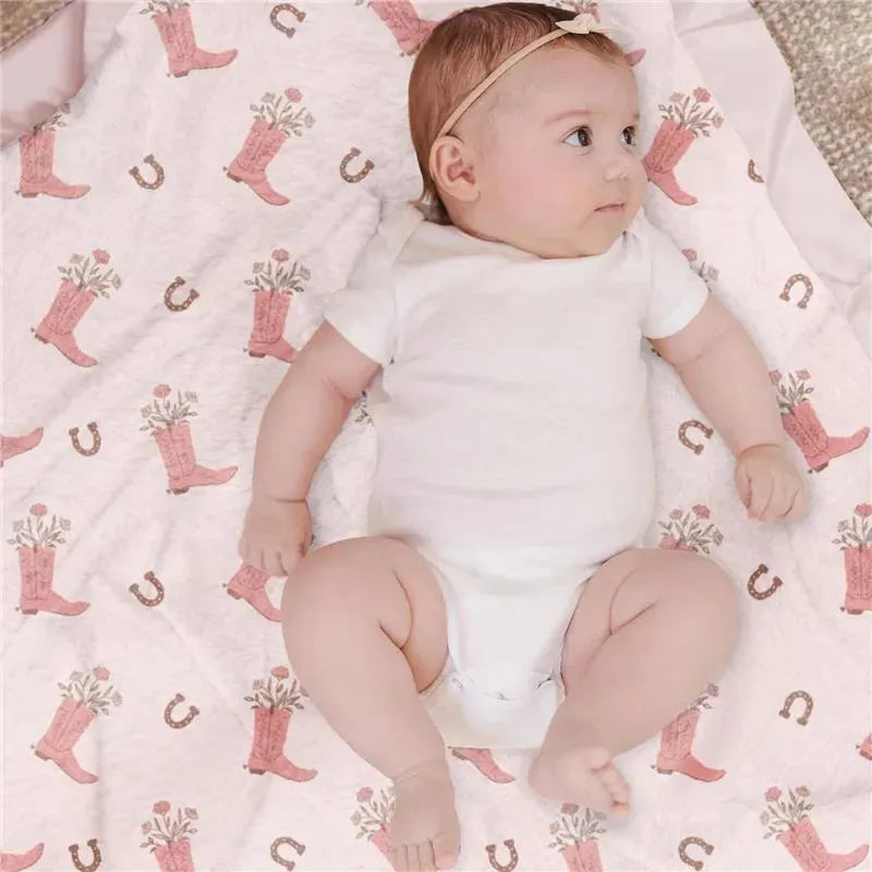 Reversible baby blanket featuring a pink cowboy boot and floral design, with a baby in a white onesie lying on it.