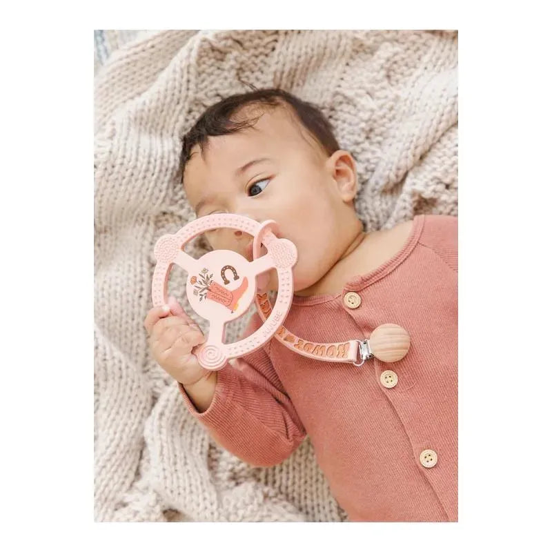 Baby holding a pink silicone teether ring with a detachable clip, featuring a floral design and a wooden button, on a knitted blanket.