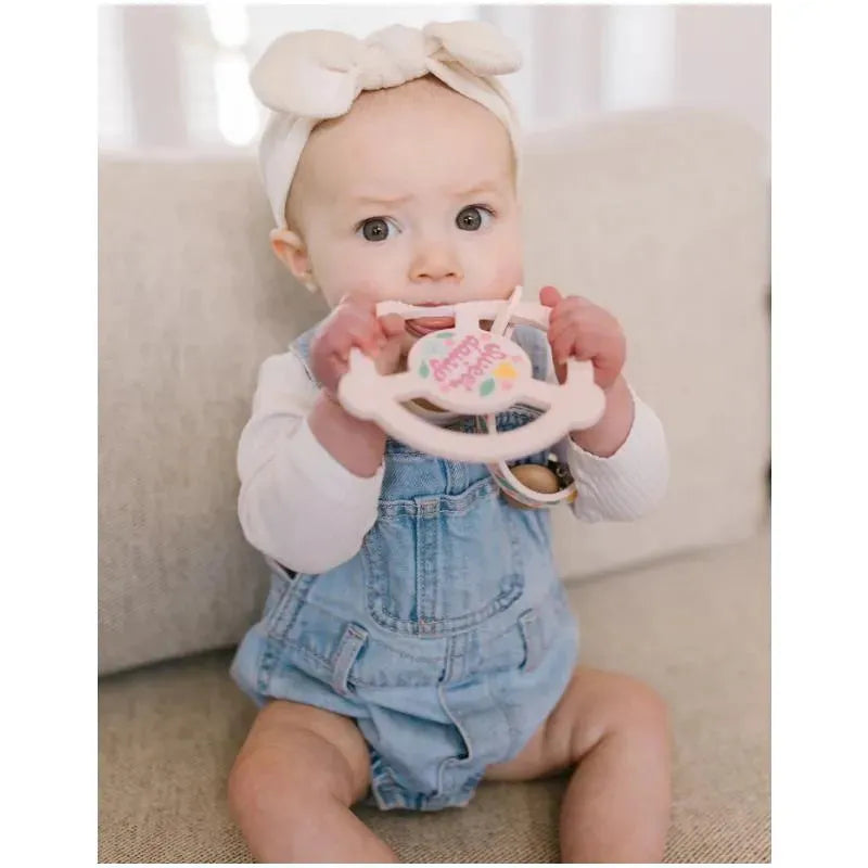 Baby sitting on a sofa, holding a pink Jujube silicone teether ring with a detachable clip, featuring a Sweet Daisy design.