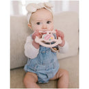 Baby sitting on a sofa, holding a pink Jujube silicone teether ring with a detachable clip, featuring a Sweet Daisy design.