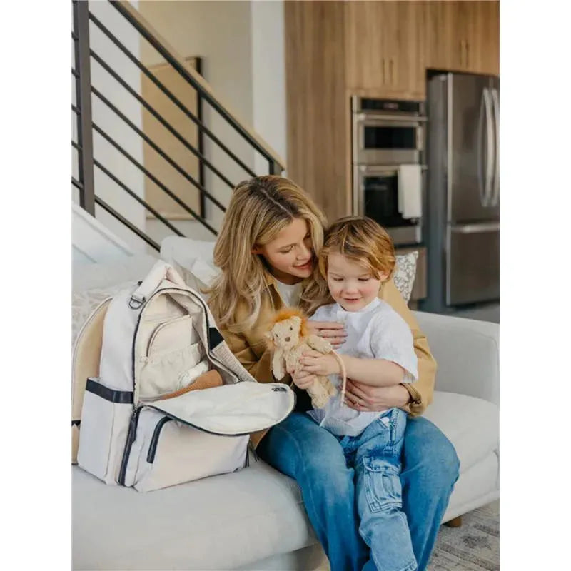A woman and a child sit together on a couch, engaging with a plush toy while a cream-colored JuJuBe x Witney Carson Classic Backpack is beside them.