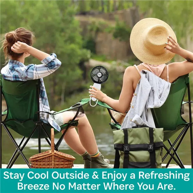 Two people sitting in camping chairs by a river, one holding a black and rose gold Kidco turbofan, promoting outdoor comfort.