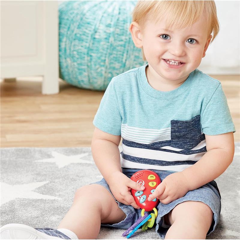 Toddler with short blond hair smiles while sitting on a rug, holding a red toy phone and colorful keys.