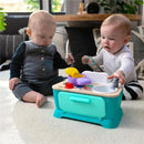 Two babies interact with the Kids II Be + Hape Magic Touch Kitchen, playfully pretending to cook with colorful utensils and a toy sink.