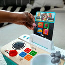 Hand placing a food card into the Kids II - Be + Hape Magic Touch Kitchen, featuring buttons and cooking elements.