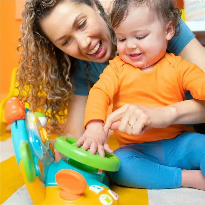 A toddler in an orange shirt plays with the Bright Starts Lights & Colors Driver Toy Steering Wheel while an adult encourages him.