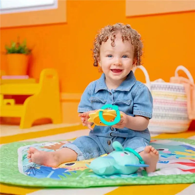 Toddler in denim outfit smiling while sitting on a colorful activity gym, holding a toy and surrounded by plush toys.