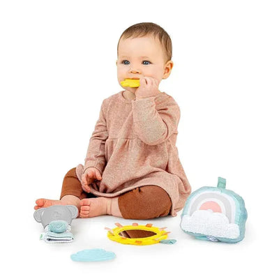 Baby sitting on a white background, holding a yellow teether, surrounded by colorful toys including an elephant, sun, cloud, and rainbow.