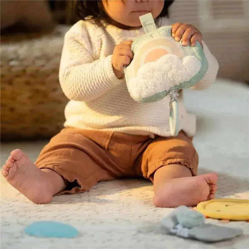 A child in a cozy sweater sits on a soft surface, holding a plush toy shaped like a cloud with a rainbow.