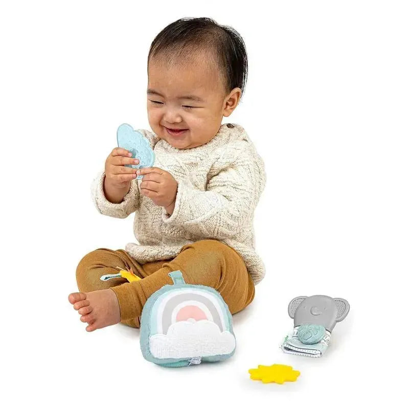 A smiling baby holds a light blue cloud-shaped toy, surrounded by colorful plush toys, including a rainbow and elephant.