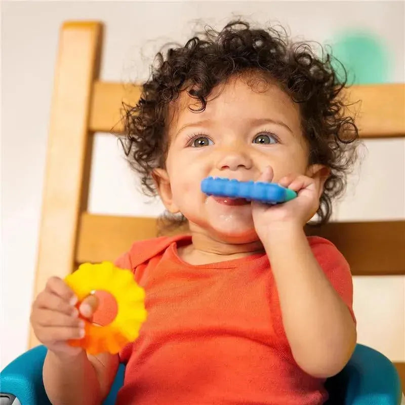 Toddler in an orange shirt chewing on a blue and green silicone teether, holding a yellow flower-shaped teether, seated in a high chair.
