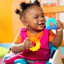 Smiling baby sitting in a high chair, holding a blue rainbow silicone teether and a yellow ring teether.